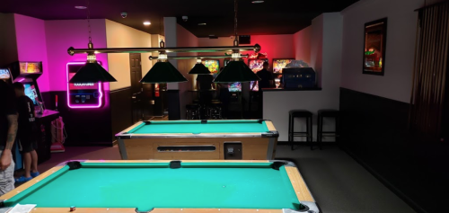A dimly lit game room featuring two pool tables, arcade machines, and neon lights in the background.