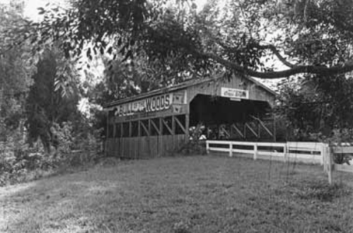 Black and white photo of a covered bridge with a sign reading "Pulaski Woods," surrounded by trees and grass.