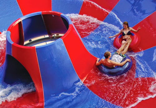 Two people on colorful inner tubes slide down a water ride with a large funnel in the center, surrounded by splashing water.