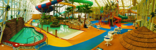 A vibrant indoor water park featuring slides, a pool, palm trees, and colorful play areas.
