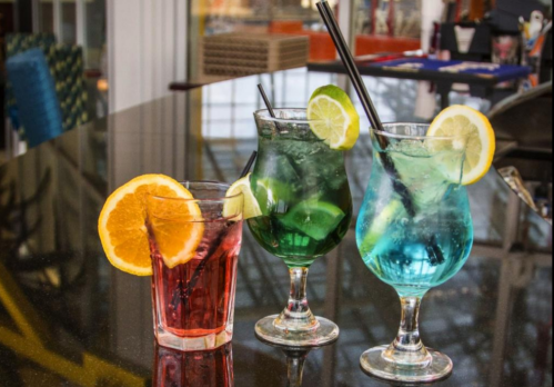 Three colorful cocktails on a table: a red drink with an orange slice, and two blue and green drinks with lime and lemon.