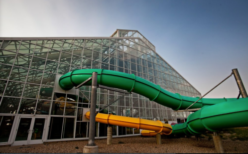 A large glass building with green and orange water slides extending from its side, set against a clear sky.