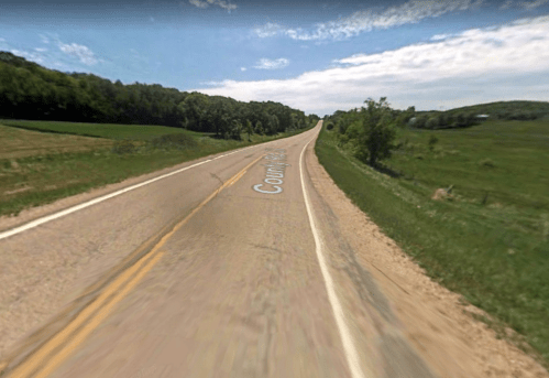 A winding country road stretches into the distance, flanked by green fields and trees under a blue sky.