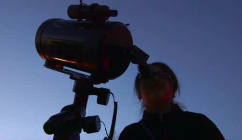A silhouette of a girl looking through a telescope against a twilight sky.