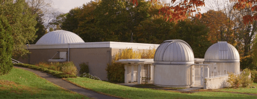 A modern observatory with two domed roofs, surrounded by trees and a pathway, in a scenic outdoor setting.