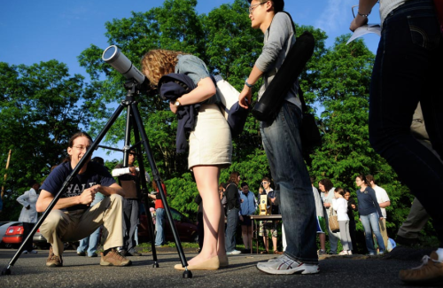 A group of people gathers outdoors, with one person looking through a telescope and others waiting in line.