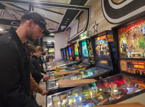 A group of people playing pinball machines in a lively arcade setting, with colorful game displays and drinks on the table.
