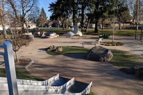 A playground with slides, climbing structures, and seating areas surrounded by trees and grassy patches.