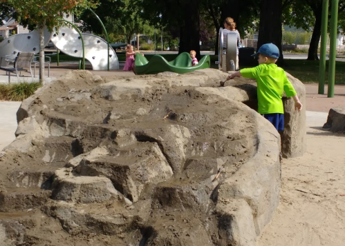 A child in a green shirt explores a sandy play structure with rocks, while other kids play in the background.