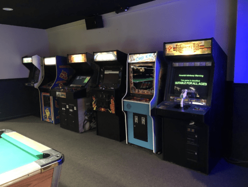 A row of vintage arcade machines next to a pool table in a dimly lit game room.