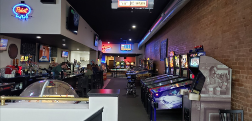 A lively bar with arcade games, pinball machines, and a cozy atmosphere, featuring neon lights and a brick wall.