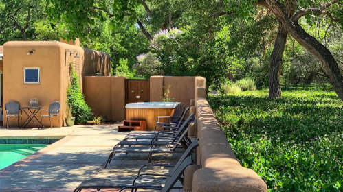 A serene outdoor space featuring a hot tub, lounge chairs, and lush greenery near a pool and adobe-style building.
