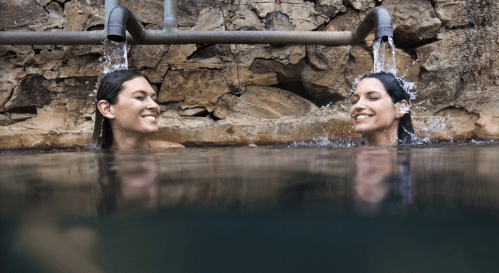 Two women smiling and enjoying a natural hot spring, with water flowing from pipes above them.