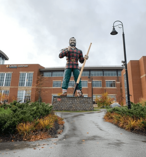 A large statue of a lumberjack in plaid, holding a log, stands in front of a modern building on a cloudy day.