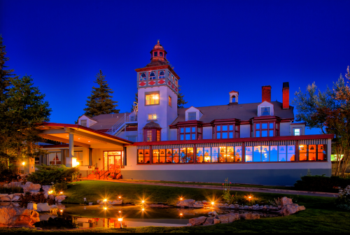 A beautifully lit historic building at dusk, featuring a tower and large windows, surrounded by a serene garden pond.