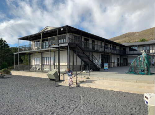 A two-story building with a staircase, surrounded by gravel and greenery, under a partly cloudy sky.