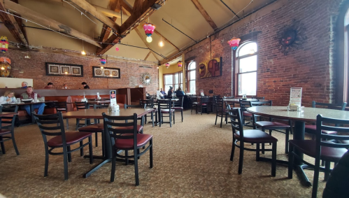 Interior of a restaurant with brick walls, empty tables, and colorful hanging lights. People are seated in the background.
