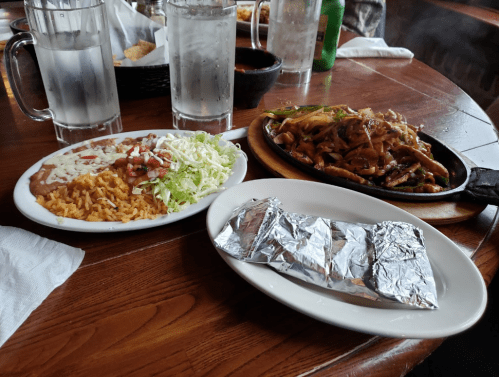 A plate with rice, beans, lettuce, and diced tomatoes, alongside sizzling fajitas wrapped in foil and two glasses of water.