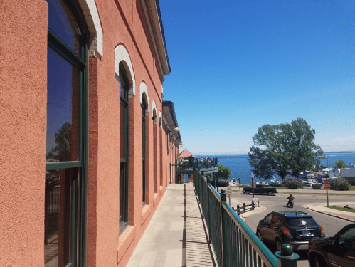 A sunny waterfront view along a pink building with a railing, leading to a marina and blue water in the distance.