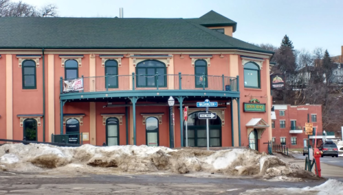 Historic building with a green roof, balcony, and signs, surrounded by snow and a street in a quaint town.