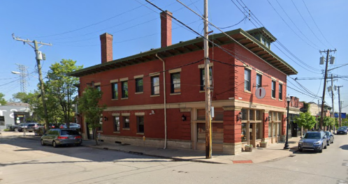 A red brick building at a street corner, featuring large windows and a green roof, with parked cars nearby.