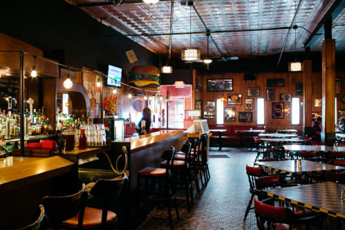 A cozy restaurant interior with a bar, wooden decor, and colorful artwork on the walls, featuring empty tables and chairs.