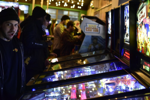 A dimly lit arcade with people playing pinball machines, featuring colorful lights and a man looking at the camera.