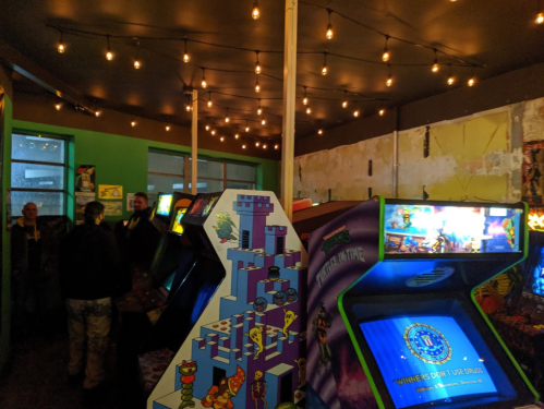 A retro arcade with colorful game machines and string lights, featuring people socializing in the background.