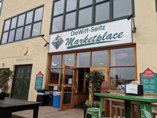 Entrance to DeWitt-Seitz Marketplace with large windows and a sign displaying hours and "Open" status.