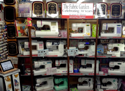 Display of sewing machines on shelves, featuring Janome models and awards for The Fabric Garden's 30th anniversary.