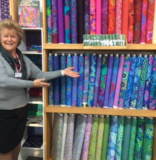 A smiling woman stands beside a display of colorful fabric rolls in a store, showcasing various patterns and colors.