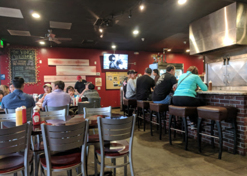 A busy restaurant interior with patrons seated at tables and a bar, featuring a TV and colorful decor on the walls.