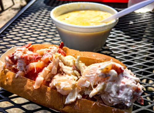 A lobster roll filled with lobster meat and coleslaw, next to a bowl of creamy corn chowder on a black table.