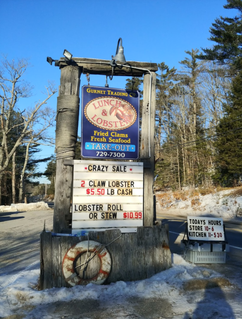 Sign for Gurnet Trading Co. featuring seafood specials: $5.50/lb lobster, $10.99 lobster roll or stew. Snowy background.