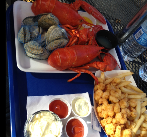 A tray with two lobsters, clams, fries, and dipping sauces, alongside a bottle of water on a table.