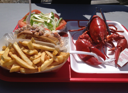 A tray with a lobster, a lobster roll, and a side of fries, accompanied by a fresh vegetable salad.