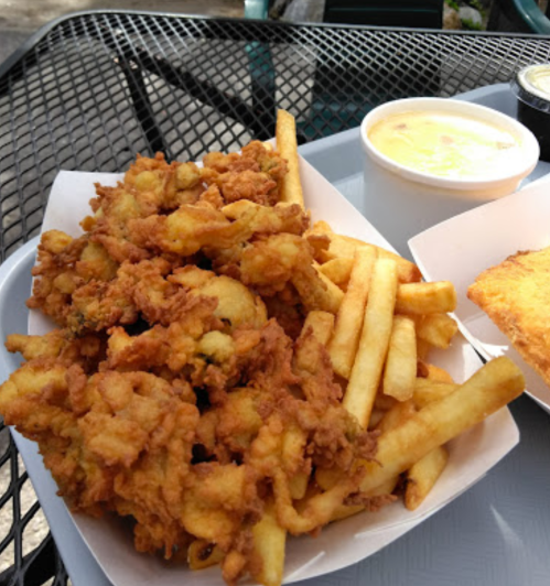 A tray of crispy fried seafood, golden fries, a cup of creamy soup, and a slice of bread on a metal table.