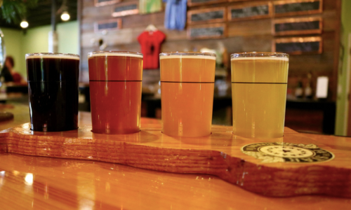Four glasses of beer in varying colors, displayed on a wooden flight board against a rustic background.