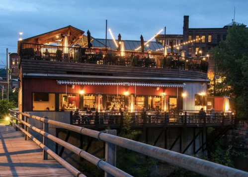 A cozy restaurant with outdoor seating, illuminated at dusk, overlooking a river and surrounded by city buildings.