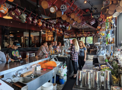 A lively bar scene with patrons at the counter, colorful mugs hanging above, and staff serving drinks.