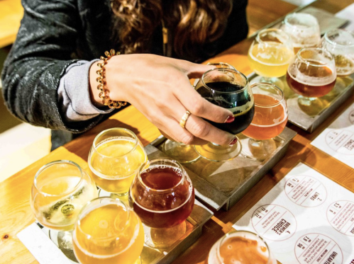 A hand reaches for a dark beer among a flight of various craft beers on a wooden table.