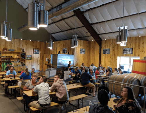 A spacious indoor venue with wooden walls, people seated at long tables, and large barrels in the background.