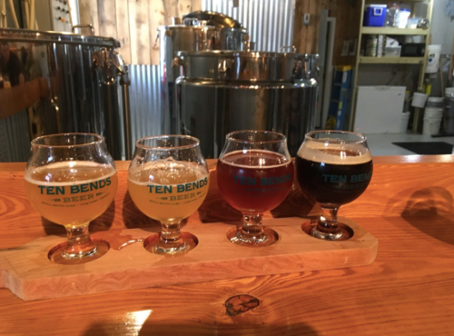 Four small glasses of beer in a wooden flight, with a brewery setup in the background.