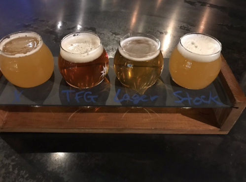 A wooden flight tray holds four small glasses of different beers, labeled with names and styles.