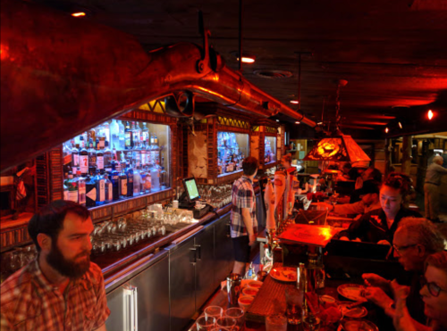 A lively bar scene with patrons, a bartender, and shelves of colorful liquor bottles illuminated by warm lighting.