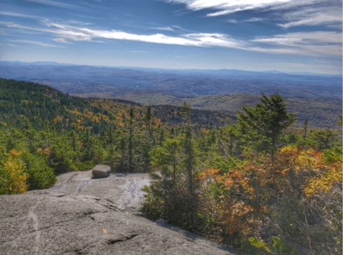 A panoramic view of colorful autumn foliage from a rocky mountain summit under a clear blue sky.