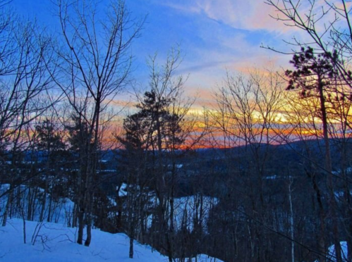 A winter landscape at sunset, featuring bare trees and a colorful sky with shades of orange, pink, and blue.