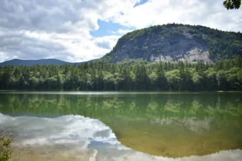 A serene lake reflects a rocky mountain and lush green trees under a partly cloudy sky.