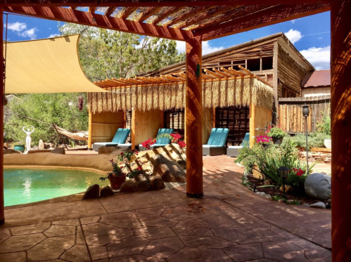 A serene outdoor space featuring a pool, lounge chairs, and lush greenery, framed by rustic architecture and a shaded pergola.