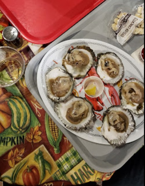 A platter of fresh oysters on a table with a drink, lemon wedge, and snacks in the background.